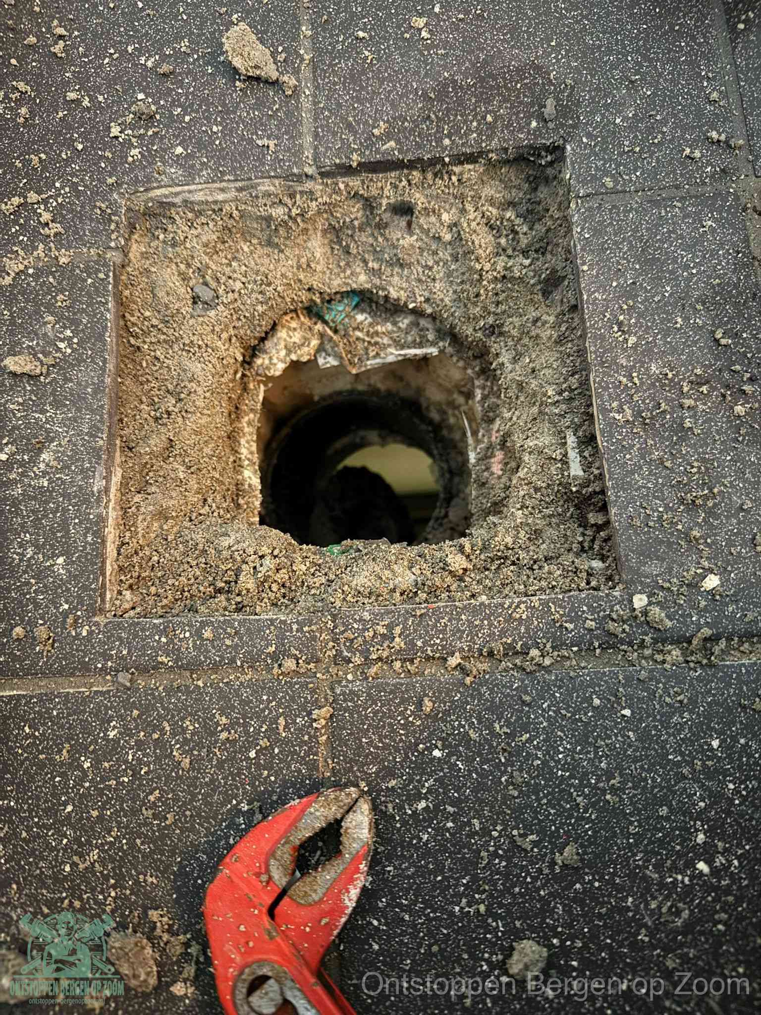 Een geopend rioolputje in een betontegelvloer met losgemaakt rooster en een zichtbare ronde afvoerbuis, het achterhalen waar de afvoerverstopping zit in Bergen op Zoom