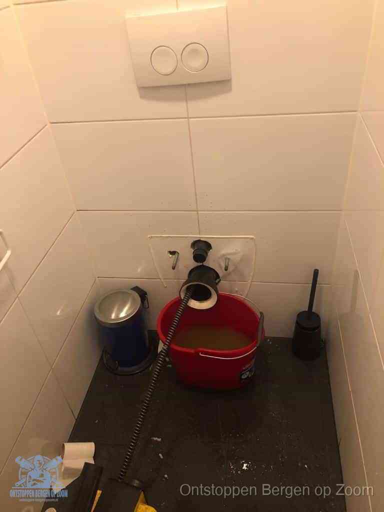 Een ontstopte wand-wc met verwijderde toiletpot waar een ontstoppingsveer in de wanduitgang verdwijnt, het achterhalen waar de verstopping zit in Bergen op Zoom