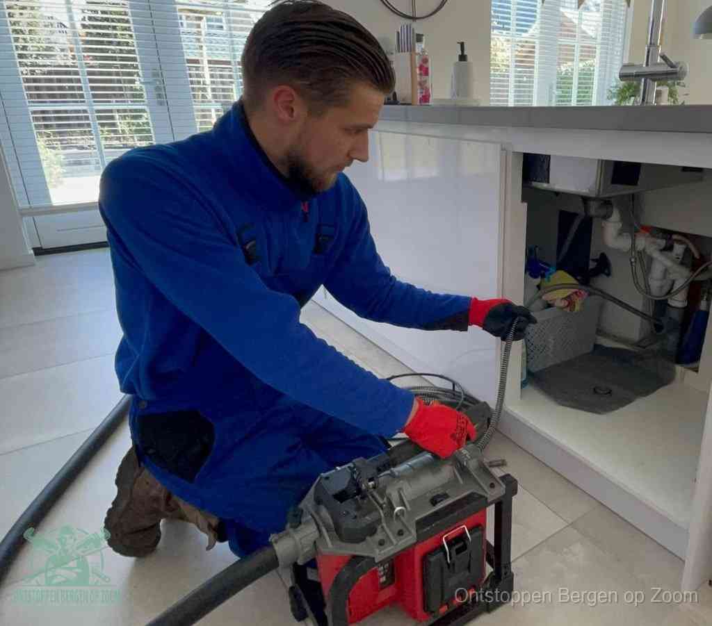 Een monteur met rode handschoenen knielt in een keuken en voert een elektrische ontstoppingsveer in tijdens het professioneel afvoer ontstoppen in Bergen op Zoom