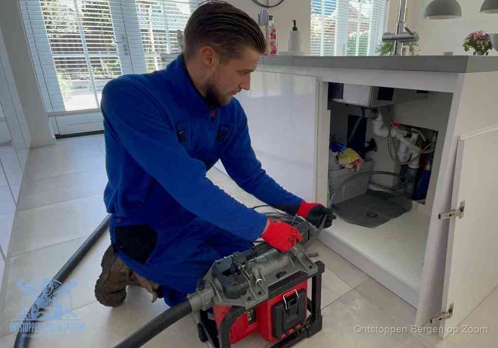 Een monteur voert een professionele elektrische ontstoppingsmachine in de afvoer onder de spoelbak tijdens het gootsteen ontstoppen in Bergen op Zoom